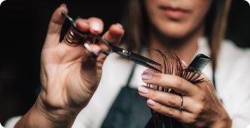 “Close-up of a hairstylist’s hands cutting hair with scissors, demonstrating precision and care in a salon setting.”